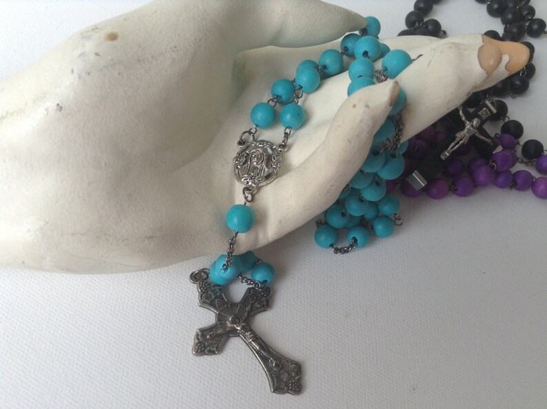Puede incluir: Un rosario azul turquesa con un crucifijo de plata y un dije de plata con una imagen de Jes&uacute;s. El rosario est&aacute; sobre una superficie blanca.