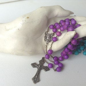 Puede incluir: Un rosario de madera morado con un crucifijo de plata y un dije de plata con una imagen de Jes&uacute;s. El rosario se muestra en una mano de maniqu&iacute; blanca.