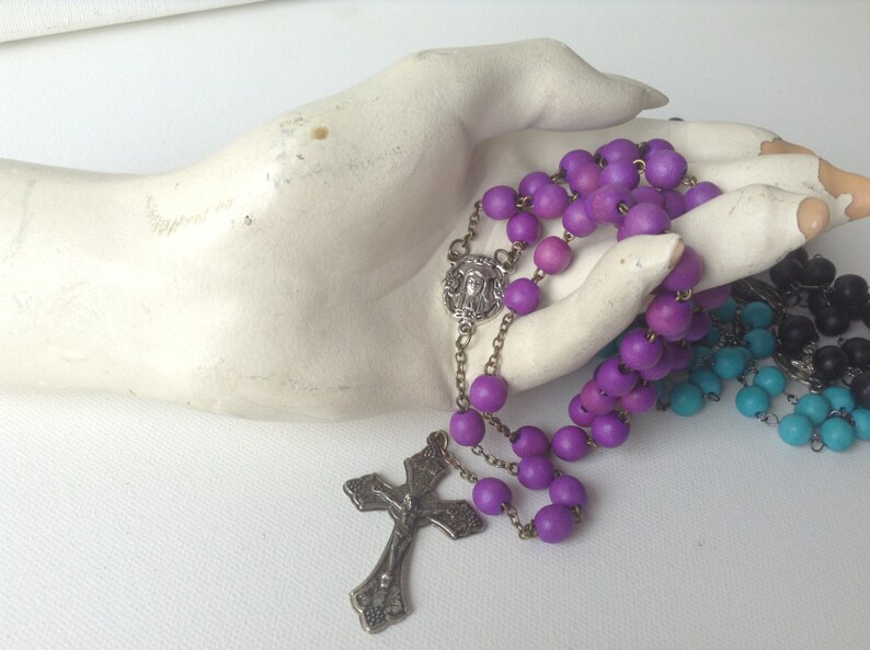 Puede incluir: Un rosario de madera morado con un crucifijo de plata y un dije de plata con una imagen de Jes&uacute;s. El rosario se muestra en una mano de maniqu&iacute; blanca.