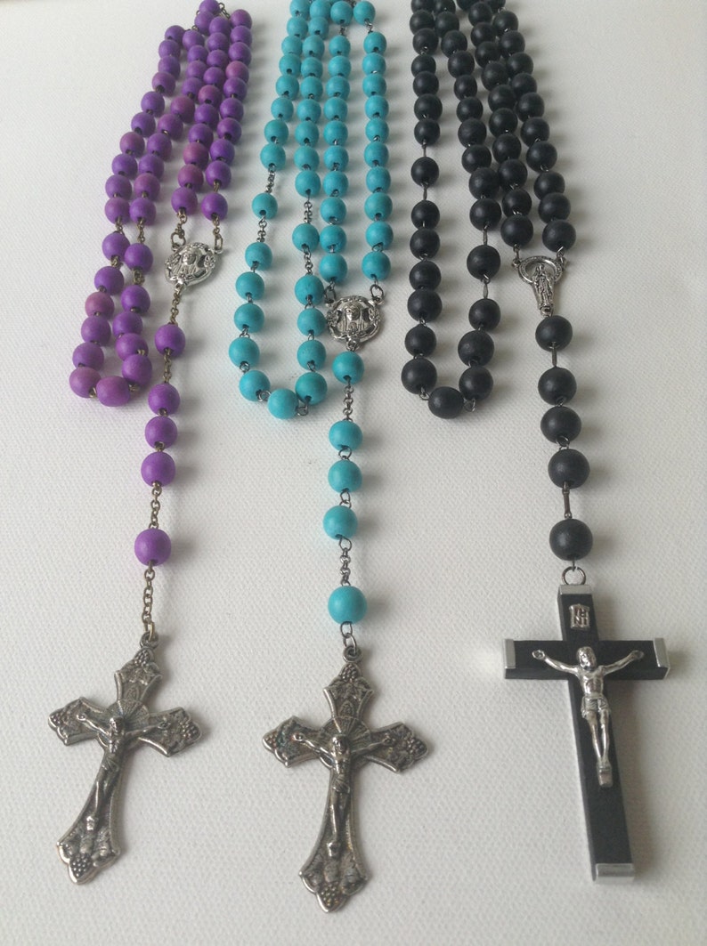 Puede incluir: Tres rosarios de madera con cuentas de diferentes colores: morado, turquesa y negro. Cada rosario tiene un crucifijo de plata y una pieza central de plata.