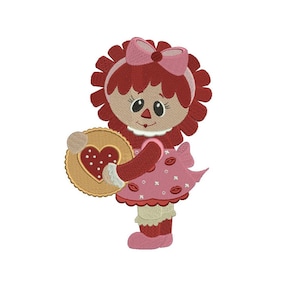 Può includere: Una bambola di pezza dai capelli rossi con un vestito rosa e un biscotto a forma di cuore. La bambola ha un fiocco rosa nei capelli e tiene il biscotto nella mano destra.