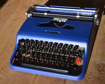 May include: A vintage blue typewriter with black keys and a red button. The typewriter has a silver carriage and the text "Lettera 22" is visible. The machine sits on a wooden surface, with sunlight casting shadows.