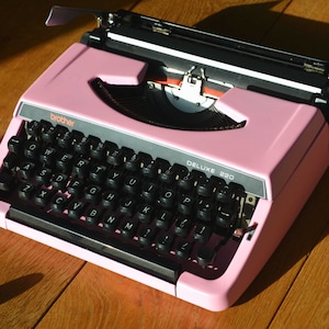 以下が含まれることがあります： 黒色のキーボードとキャリッジリターンレバーが付いたピンク色のブラザーデラックス220タイプライター。タイプライターは黒と白のリボンを使用しています。