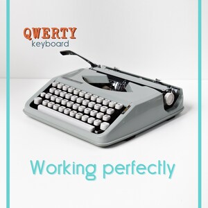 Puede incluir: Una máquina de escribir gris vintage con teclas blancas. La imagen incluye el texto "QWERTY keyboard" en naranja y "Working perfectly" en azul. La máquina de escribir está sobre un fondo blanco.