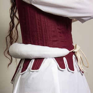 Könnte beinhalten: Ein weinrotes Korsett mit vertikalen Nähten und weißer Bordüre. Das Korsett wird über einer weißen Bluse und einem Rock getragen. Der untere Rand des Korsetts hat ein gewelltes Design. Eine kissenartige Form in Weiß wird um die Taille getragen.