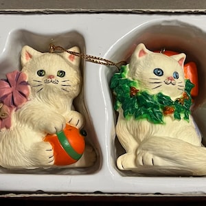 May include: Two cream-coloured cat ornaments. One cat holds an orange ball with green stripes and a pink bow. The other cat wears a green wreath with red berries. Both have blue eyes and black whiskers. They are in a white tray.