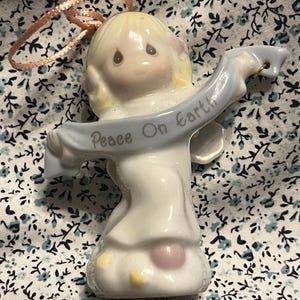 May include: A ceramic angel figurine with a light blue banner that reads "Peace On Earth." The angel has blonde hair, a white dress, and pink accents. A pink ribbon is attached for hanging. The figurine is on a floral patterned background.