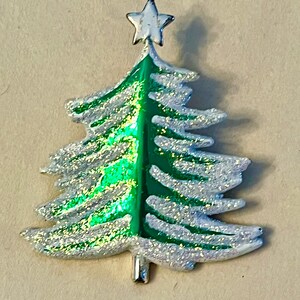 May include: A Christmas tree brooch with a silver glitter outline and a green centre. A silver star sits atop the tree. The brooch has a silver pin on the back.