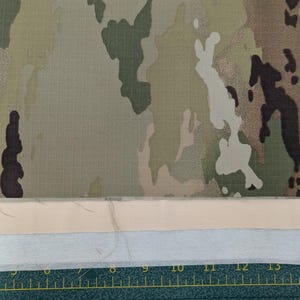 May include: Close-up of a camouflage fabric with a beige and white fabric underneath. A ruler is visible at the bottom of the image.