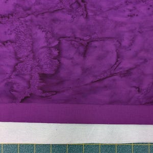 May include: Purple fabric with a marbled pattern. The fabric has a purple border and is layered on top of a white fabric.