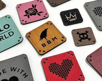 Op de afbeelding: Een verzameling vierkante en rechthoekige kunstlederen labels in diverse kleuren zoals rood, roze, bruin, grijs en groenblauw. Elk label is voorzien van voorgeponste hoekgaten en lasergegraveerde ontwerpen zoals harten, schedels en tekst.