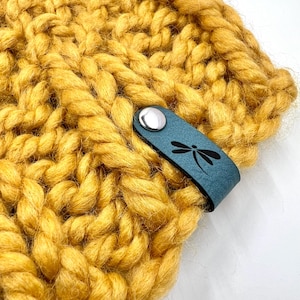 以下が含まれることがあります： マスタードイエローのニット帽に、トンボのデザインが施された青い革のストラップが付いている。