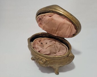 Könnte beinhalten: Eine ovale Schmuckschatulle in Goldfarbe mit einem rosafarbenen Samt-Innenraum. Die Schachtel hat drei Beine und aufwendige Blumendetails.