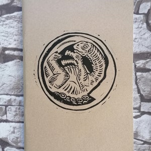 Peut inclure: Un carnet en papier brun avec une illustration en noir et blanc d'une créature avec des tentacules. L'illustration est dans un cercle.