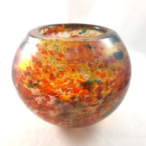 Puede incluir: Un jarrón de vidrio redondo con un patrón moteado en tonos de naranja, amarillo, rosa y verde. El jarrón es transparente y tiene un acabado liso y pulido.