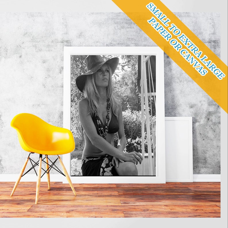 May include: Black and white photograph of a woman wearing a wide-brimmed hat and a bikini top. She is sitting on a wooden bench with a leafy background.