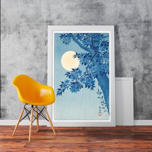 Puede incluir: Una impresión japonesa en madera grabada enmarcada que representa un árbol con flores azules y una luna llena en el fondo. La impresión está en un marco blanco y está colgada en una pared gris.
