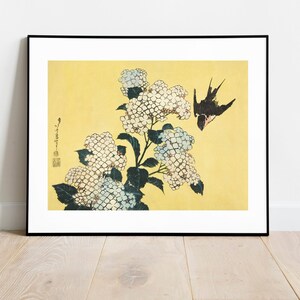 Puede incluir: Una impresión japonesa vintage en madera que representa una flor de hortensia blanca con una golondrina negra en vuelo sobre un fondo amarillo. La impresión está enmarcada en negro.