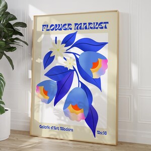 Puede incluir: Un póster con el texto "FLOWER MARKET" en una fuente retro. El póster presenta una ilustración estilizada de una rama de flores azules con flores blancas y acentos coloridos. El texto "Galerie d'Art Moderne" y "No.18" se encuentra en la parte inferior del póster.