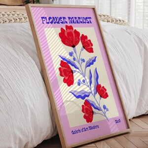 Puede incluir: Un póster enmarcado con fondo rosa y un patrón de rayas blancas y beige. El póster presenta una ilustración estilizada de flores rojas con hojas azules y el texto "FLOWER MARKET" en una fuente retro. El texto "Galerie d'Art Modern" y "No.3" también están incluidos en el póster.