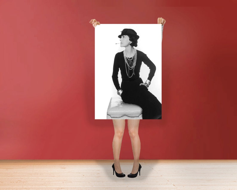 以下が含まれることがあります： 帽子、長袖トップス、スカートを着た女性の白黒ポートレート。スツールに座っています。彼女は複数の真珠のネックレス、時計、ブレスレットを身に着けています。口にはタバコがあります。背景は無地の赤です。