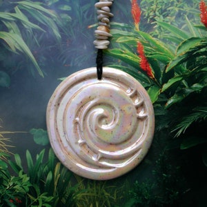 May include: A white ceramic pendant with a swirling design, hanging from a string of white beads. The pendant is iridescent and has a subtle sparkle. The background is a blurry image of tropical plants.