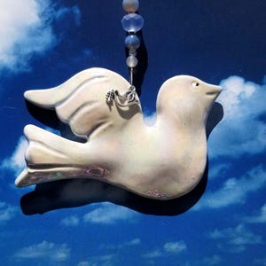 May include: A white ceramic dove ornament with iridescent glitter, hanging from a blue and white beaded string. The dove has a small silver charm attached to its wing.