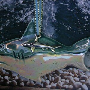 May include: A ceramic ornament shaped like a shark with a smaller shark ornament hanging from its back. The larger shark is green and iridescent, while the smaller shark is silver. The ornament is hanging from a gold chain.