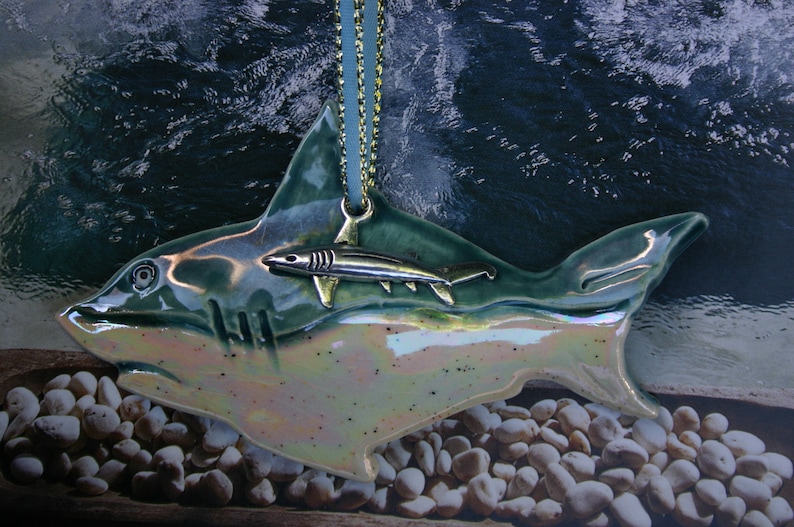 May include: A ceramic ornament shaped like a shark with a smaller shark ornament hanging from its back. The larger shark is green and iridescent, while the smaller shark is silver. The ornament is hanging from a gold chain.