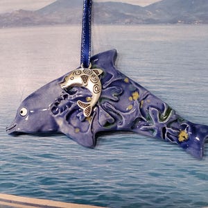 May include: A ceramic dolphin ornament in shades of blue, with a smaller silver dolphin charm attached. The ornament has a blue ribbon for hanging and is decorated with yellow accents.