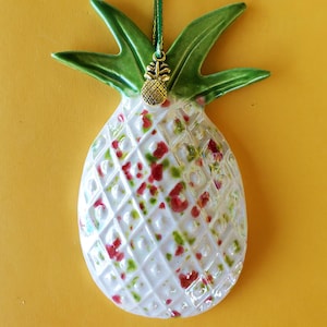 Puede incluir: Un adorno de cerámica con forma de piña que presenta una base blanca con textura de rombos, acentuada por un esmalte moteado en rojo y verde. La parte superior tiene hojas verdes y una cinta verde con un pequeño dije de piña metálico dorado.