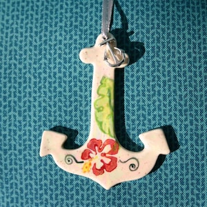 May include: Cream-coloured anchor ornament with a silver metal anchor charm and a grey ribbon for hanging. The anchor is decorated with a red and yellow hibiscus flower and green leaves. The ornament is likely ceramic or porcelain.