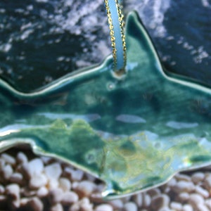 May include: A teal blue ceramic shark ornament with a gold string for hanging. The shark is detailed with a textured surface and a glossy finish.
