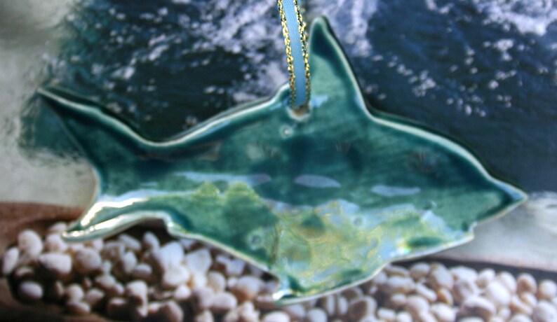 May include: A teal blue ceramic shark ornament with a gold string for hanging. The shark is detailed with a textured surface and a glossy finish.