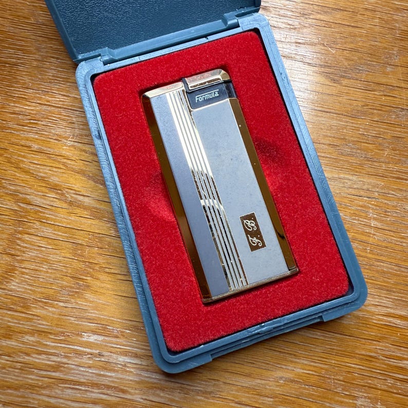 May include: A vintage silver and gold Formula lighter in a blue case with a red velvet interior. The lighter has vertical gold stripes and the initials "J.B." are visible. The case is open, revealing the lighter.