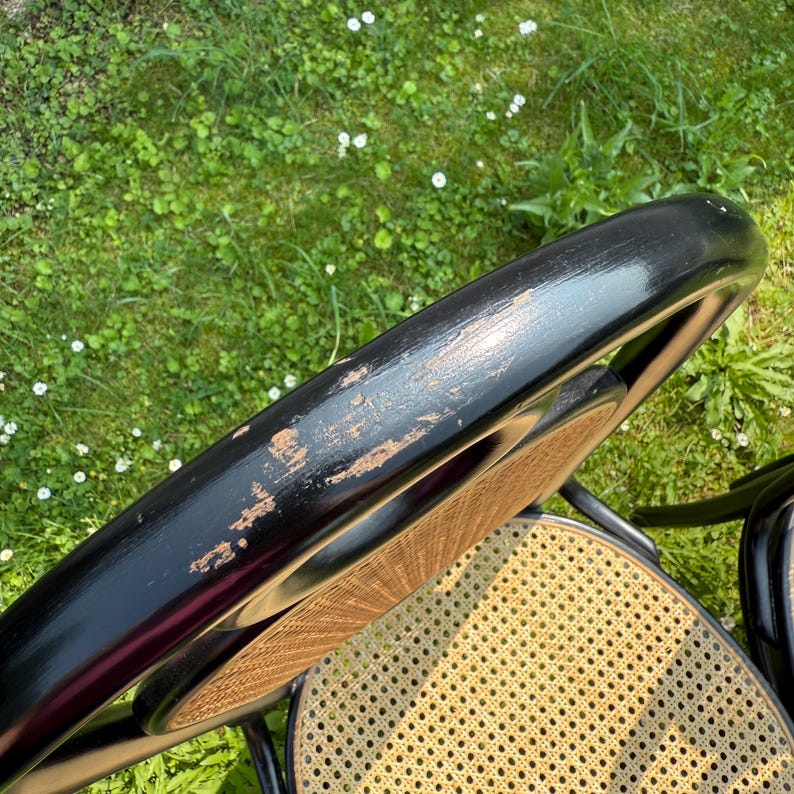 May include: Close-up of a black wooden chair with a woven cane seat. The chair's back has a curved design with visible wear and tear on the black paint, revealing the wood underneath. The chair is set against a backdrop of green grass.