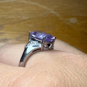 May include: Silver ring with a large, oval-cut purple gemstone. The ring has a detailed setting with smaller accent stones on the shoulders. The gemstone is the focal point, showcasing its colour and clarity.