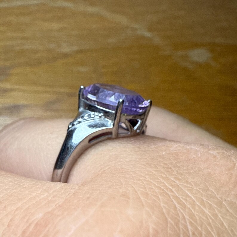 May include: Silver ring with a large, oval-cut purple gemstone. The ring has a detailed setting with smaller accent stones on the shoulders. The gemstone is the focal point, showcasing its colour and clarity.