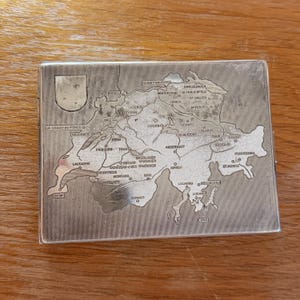 以下が含まれることがあります： スイスの詳細な地図が刻まれた銀色のシガレットケース。地図には、チューリッヒ、ジュネーブ、ベルンなどの都市名が含まれています。ケースは長方形で、左上に小さなシールドエンブレムが付いています。