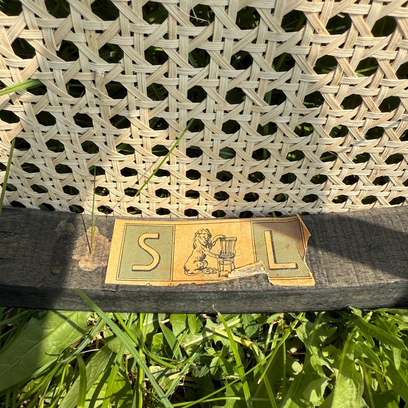 May include: Close-up of a vintage chair back with woven cane detailing. The chair's frame is dark, and a faded label with the letters "S" and "L" is visible. The label also features a lion holding a chair.