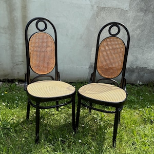 May include: Two black chairs with woven cane seats and backs. The backs have a circular design at the top. The chairs are set on a grassy surface with a concrete wall in the background.