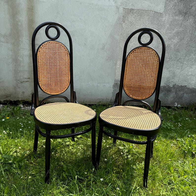 May include: Two black chairs with woven cane seats and backs. The backs have a circular design at the top. The chairs are set on a grassy surface with a concrete wall in the background.