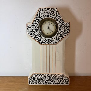 Puede incluir: Reloj de sobremesa crema con volutas negras y detalles dorados. La esfera del reloj es redonda con un marco color latón y manecillas negras. Líneas verticales doradas adornan el cuerpo del reloj, y la base presenta volutas a juego.