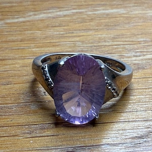 May include: A silver ring with a large, oval-cut, faceted amethyst gemstone. The ring has a detailed band with small accent stones on each side of the amethyst. The gemstone is a vibrant purple colour.