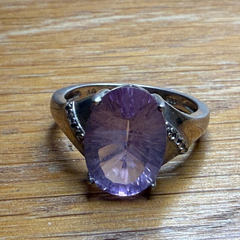 May include: A silver ring with a large, oval-cut, faceted amethyst gemstone. The ring has a detailed band with small accent stones on each side of the amethyst. The gemstone is a vibrant purple colour.