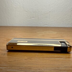 May include: A vintage gold and gray lighter with a rectangular shape. The lighter has gold trim and a black accent on one end. The top features two thin gold stripes and a small label with text. The lighter is on a wooden surface.