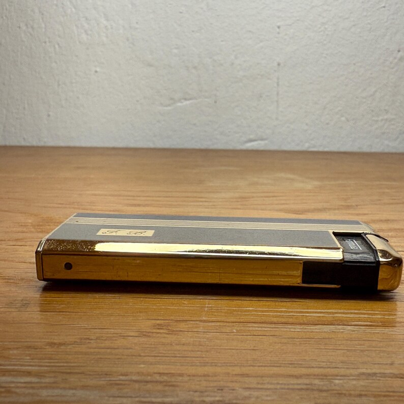 May include: A vintage gold and gray lighter with a rectangular shape. The lighter has gold trim and a black accent on one end. The top features two thin gold stripes and a small label with text. The lighter is on a wooden surface.