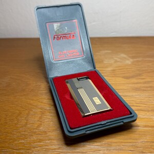 May include: A vintage Formula electronic gas lighter in a gray case with red velvet lining. The lighter is silver and gold with vertical stripes. The case lid has the word "Formula" in red. The text "Electronic Gas Lighter" is also visible.