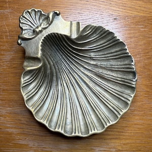 May include: A brass seashell-shaped ashtray with a textured surface and a small notch for holding items. The ashtray has a detailed, ridged design and is set against a wooden background.