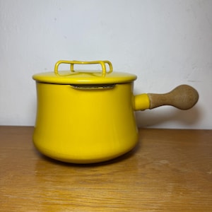 Può includere: Una casseruola in smalto giallo brillante con coperchio e manico in legno. Il manico è attaccato al lato della pentola. Il coperchio ha una piccola maniglia. La casseruola è su una superficie di legno.